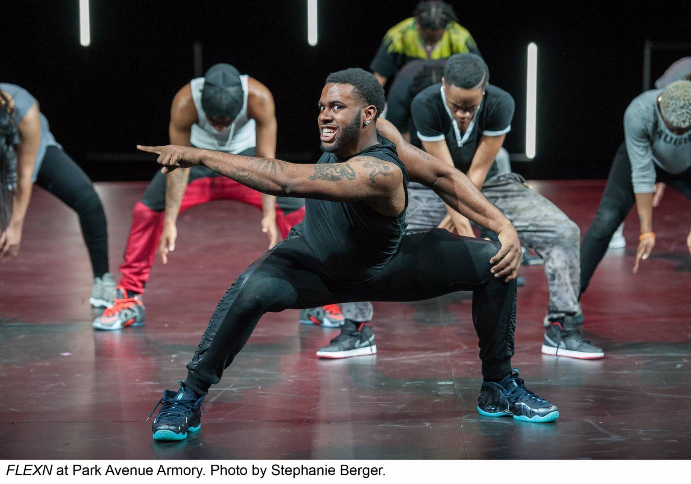 Tyme (Glendon Charles) is in the foreground wearing a menacing grin while squatting and pointing. A line of FLEXN dancers are behind him in a line also in deep squats